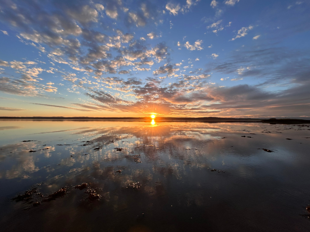 Sunset over a calm body of water. The sky is filled with a dramatic display of colourful clouds, ranging from oranges and pinks near the horizon to blues and purples higher up. The sun is setting directly in the centre of the horizon, its light reflected perfectly on the still water, creating a mirror-like effect of the sky. The water itself appears relatively shallow, with patches of seaweed or other organic matter visible near the surface. The overall scene is peaceful and serene, showcasing the beauty of a natural, tranquil sunset.