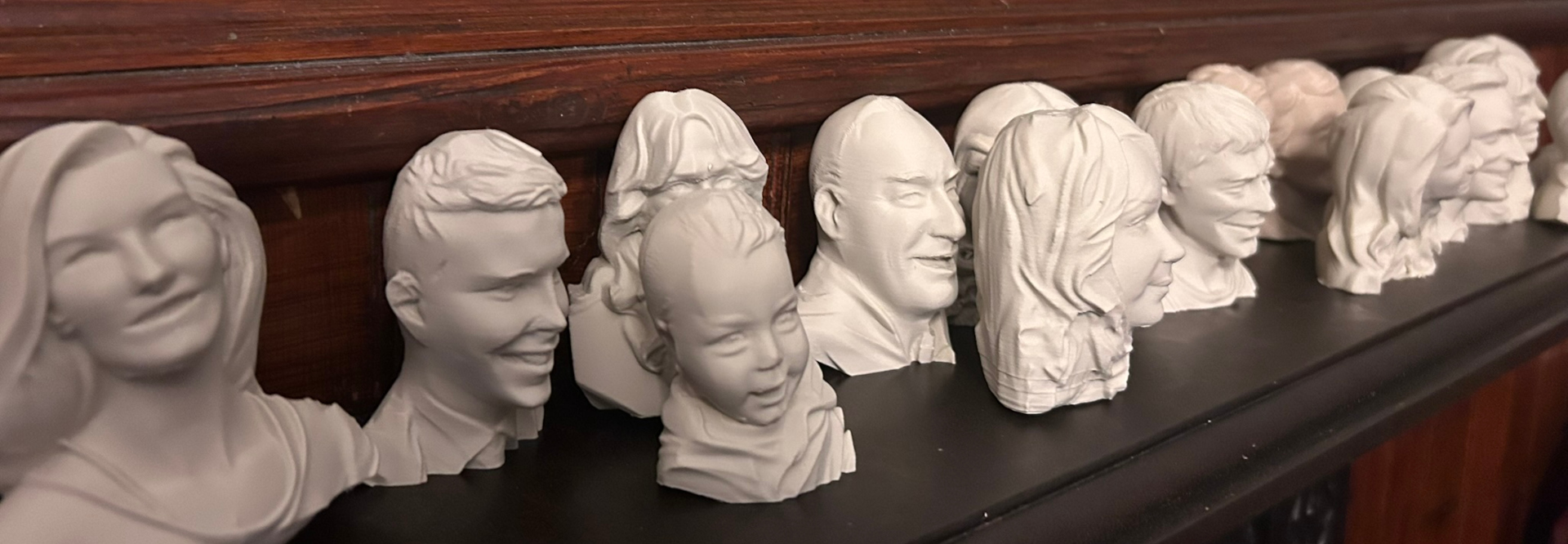 A row of small, white, three-dimensional busts, seemingly sculpted from a material like plaster or resin. They appear to be portrait busts of different individuals, likely family members, of varying ages, from a baby to adults. The busts are arranged closely together on a dark shelf or mantelpiece, creating a cohesive family portrait in three dimensions. The style is simple and realistic, focusing on capturing the likeness of each person.