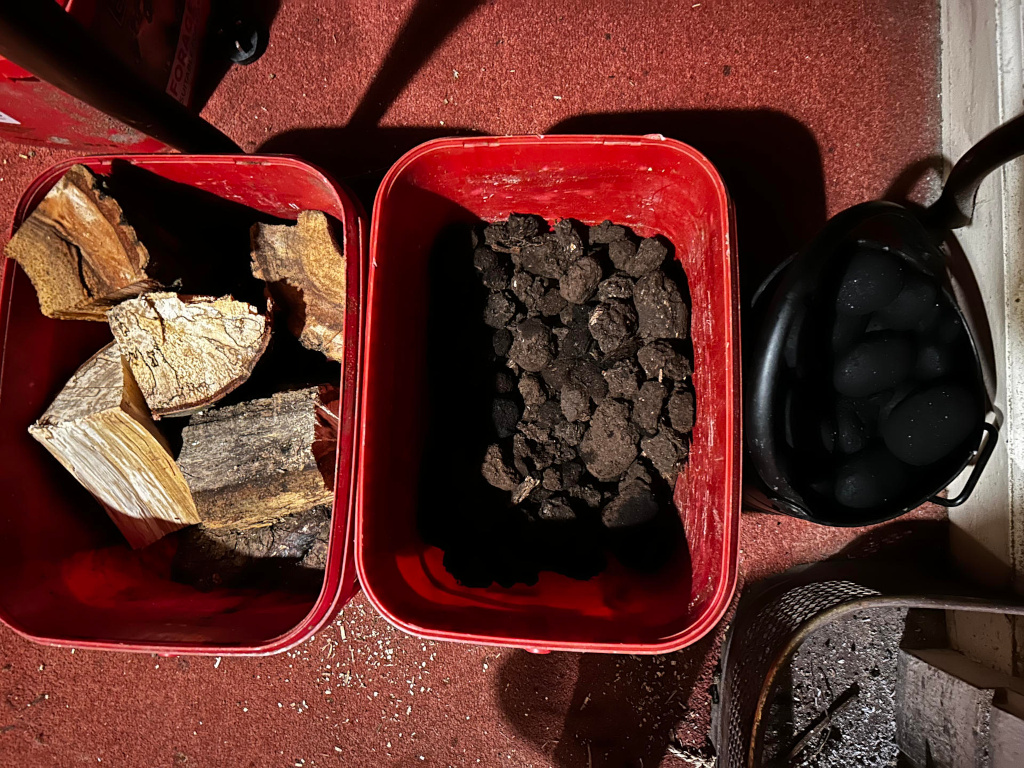 Two bright red plastic tubs positioned side-by-side on a dark reddish-brown floor. The tub on the left contains a pile of split firewood. The wood pieces vary in size and shade; some are light beige-brown, others are darker, showing varying degrees of dryness and weathering. The wood is haphazardly stacked, giving a sense of casual storage. The tub on the right is filled with a large quantity of dark brown, almost black, irregularly shaped pellets or clumps. They appear moist and earthy, possibly compost or peat. Next to the red tubs, partially obscured by the shadows and slightly out of focus, is a black metal bucket containing smooth, dark grey or black, roughly spherical objects—possibly stones or coal. Part of what looks like a metal grate or firebox is visible in the lower right corner, strewn with dark grey ash or dust.