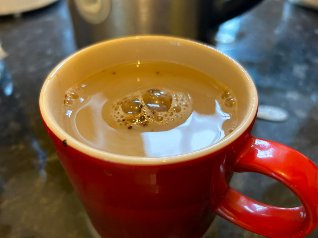 Vibrant red mug, approximately two-thirds full of a light brown liquid, which appears to be coffee or tea. The liquid shows a subtle, almost translucent quality, with a cluster of small, glistening bubbles gathered near the center, indicating recent pouring or stirring. The mug's handle, a bold and shiny red, curves gracefully to the right, inviting the viewer's eye to follow its form. The mug sits on a dark, mottled surface, suggesting a counter top or table.