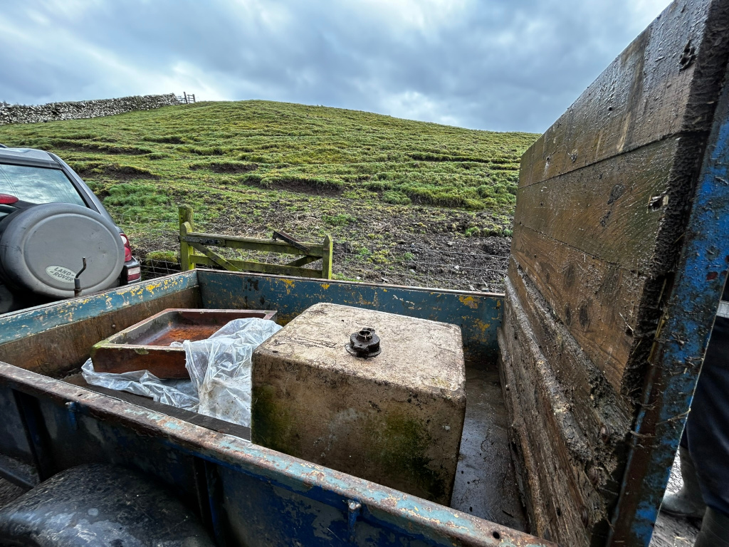 Weathered, blue metal trailer, showing signs of rust and wear, dominates the foreground. Its tailgate is partially raised, revealing its interior. The trailer bed is dirty and shows evidence of mud and water stains. Two large, rectangular concrete blocks sit in the trailer bed. The larger one is light grayish-tan, showing considerable staining and weathering, almost moss-like in texture in places. A small, dark metallic fitting is visible on its top. The smaller block appears to be a worn, terracotta-coloured trough or basin, partially covered by a translucent plastic sheet.