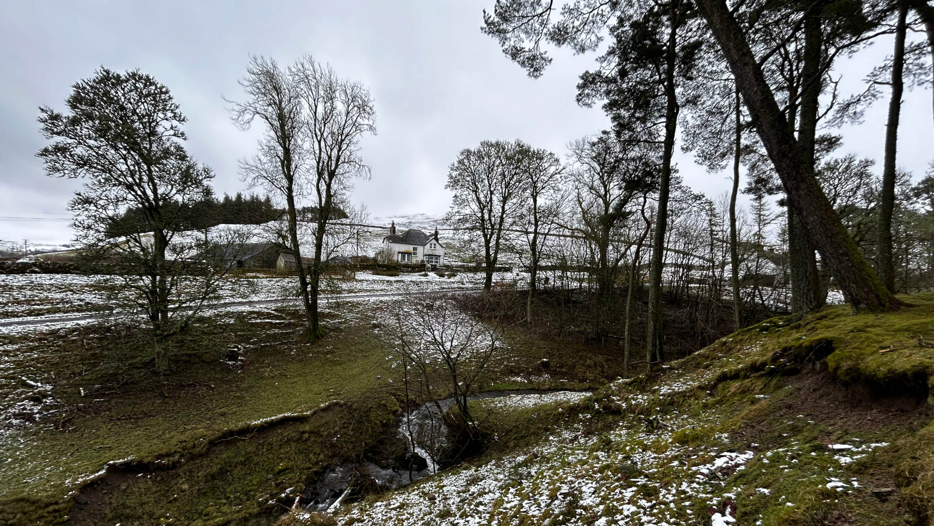 A snow-dusted landscape in the Scottish Highlands. A small stream flows through the foreground, which is covered in patchy snow and moss. Beyond the stream, the land gently slopes upward toward a cluster of leafless trees surrounding a white farmhouse and outbuildings. In the background, a darker line of distant trees and a cloudy sky complete the scene. The overall feel is one of quiet solitude and a serene, wintry atmosphere.