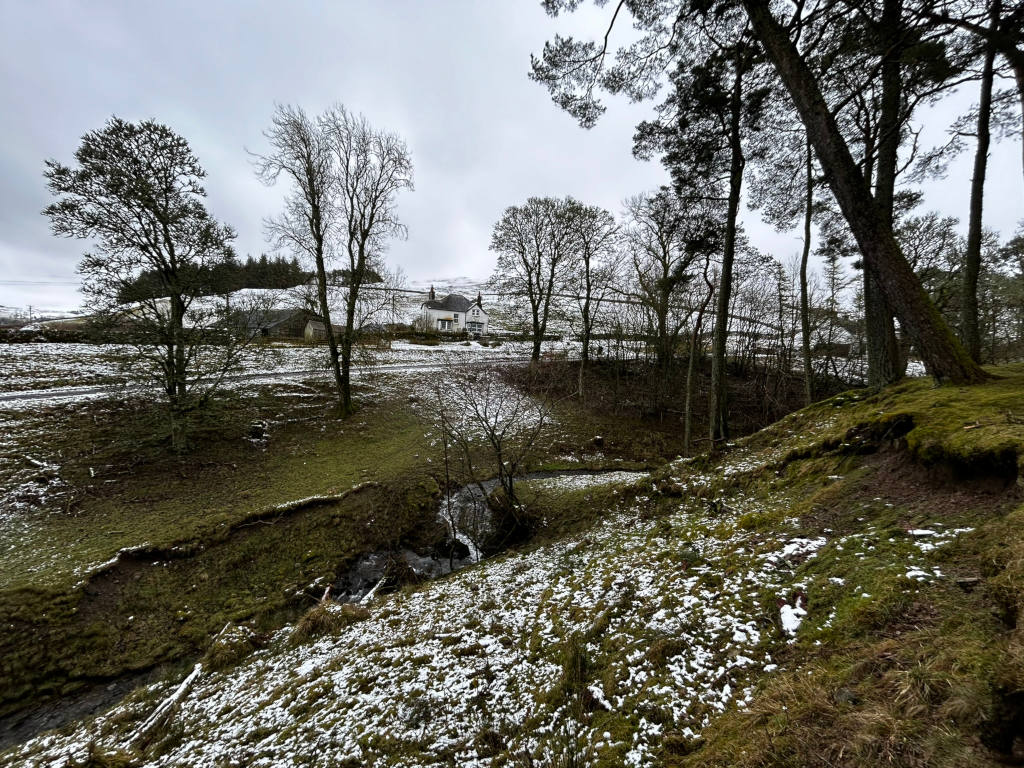 Snow-dusted landscape in the Scottish Highlands. A small stream flows through the foreground, which is covered in patchy snow and moss. Beyond the stream, the land gently slopes upward toward a cluster of leafless trees surrounding a white farmhouse and outbuildings. In the background, a darker line of distant trees and a cloudy sky complete the scene. The overall feel is one of quiet solitude and a serene, wintry atmosphere.