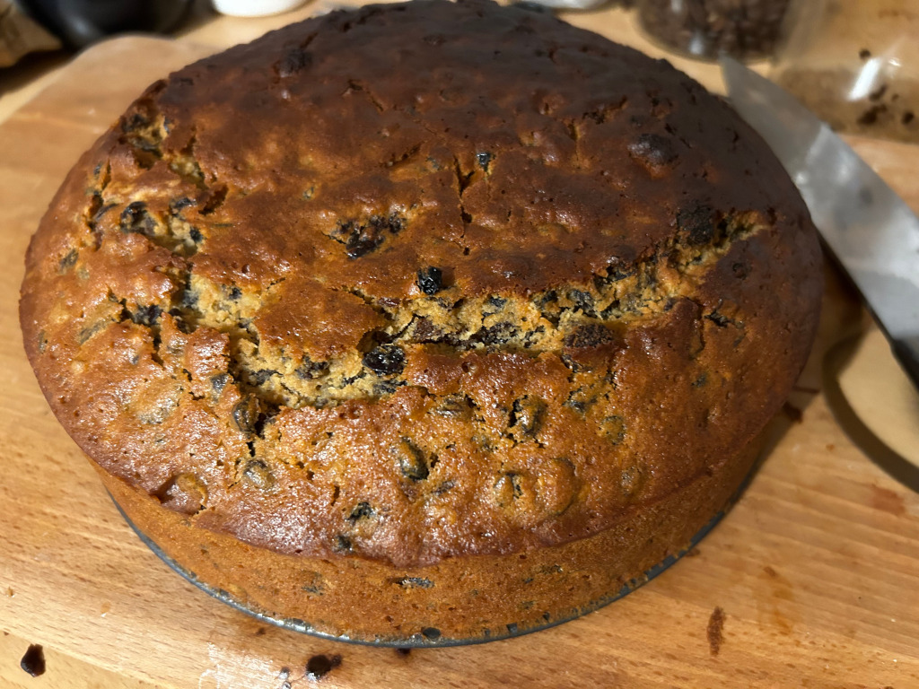 Round fruitcake, baked in a dark-coloured pan, sitting on a light brown wooden cutting board. A knife is partially visible in the upper right corner. The cake is golden brown and studded with numerous dark-coloured fruits, possibly raisins or currants. The overall appearance suggests a homemade, rustic style.