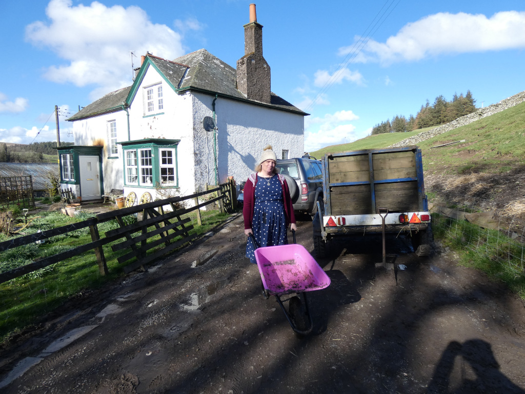 Leonie standing in front of a white house in the countryside. She's pushing a pink wheelbarrow. Behind her is a small, dark-colored vehicle towing a trailer. The scene is set in a rural area with a grassy hillside in the background. The overall impression is one of rural life and perhaps gardening.