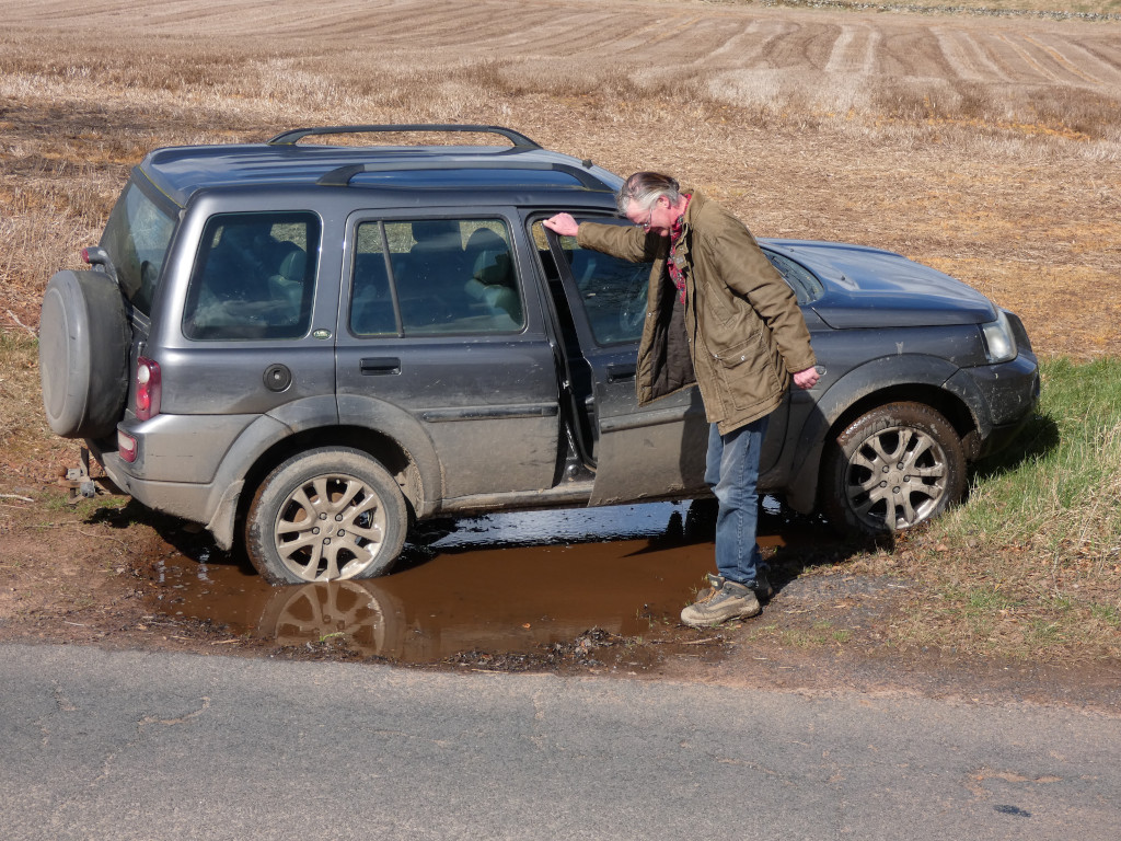 Dark grey Land Rover parked next to a muddy puddle on a rural road. Charlie attempting to step into the vehicle, appearing to assess the situation. The background features a ploughed field under a bright, sunny sky. The overall impression is one of a minor, everyday inconvenience in a rustic setting.
