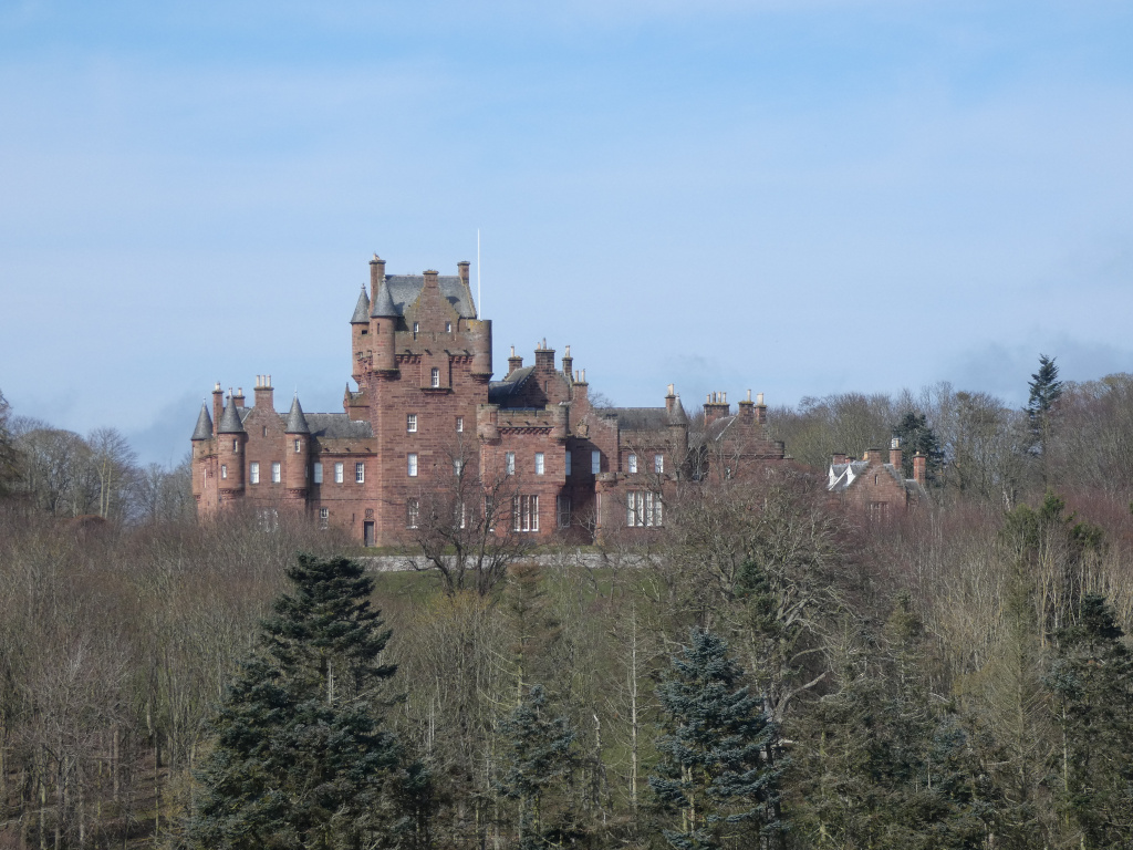 Large, red sandstone castle perched atop a hill, partially obscured by a foreground of leafless trees. The castle is multi-turreted, exhibiting a blend of architectural styles suggestive of a Scottish baronial castle. The sky is a clear, pale blue.