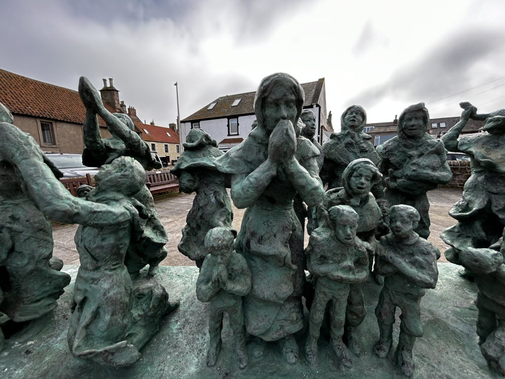 Bronze sculpture depicting a group of people, predominantly women and children, appearing to be in a state of distress or prayer. The figures are clustered together, their postures suggesting grief, supplication, or perhaps a desperate exodus. The style is realistic, yet imbued with a sense of emotional weight. The background features a typical village setting, suggesting a historical or narrative context. The overall impression is one of hardship and collective suffering.