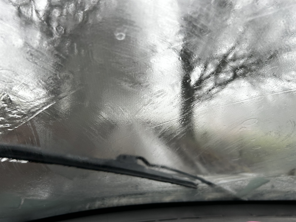 View from inside a car, looking out through a rain-streaked windshield. The wipers are visible, and the blurred background suggests a rainy day with trees visible through the water droplets. The overall effect is one of blurring and indistinctness, suggestive of a journey on a stormy day.
