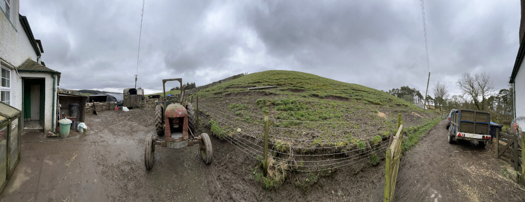 Panoramic view shows a muddy farmyard scene on an overcast day. A vintage tractor is parked near a farmhouse, with various farm implements and structures visible. A grassy hill slopes upward, enclosed by a wire fence. A trailer is parked further down the muddy track. 
