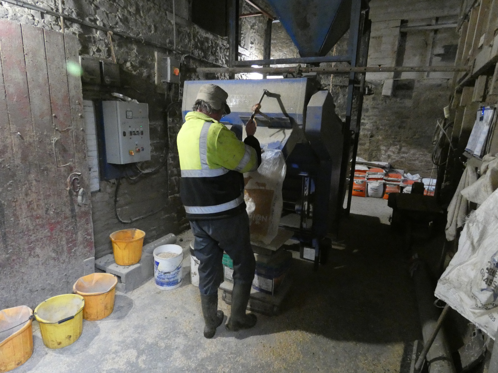 Charlie in a high-visibility vest operating a grain processing machine in what appears to be a basement or cellar. He is filling a large bag with grain or a similar product from the machine. The setting is rustic and industrial, with old stone walls, buckets, and other equipment visible. The overall impression is one of manual labour in a traditional setting.
