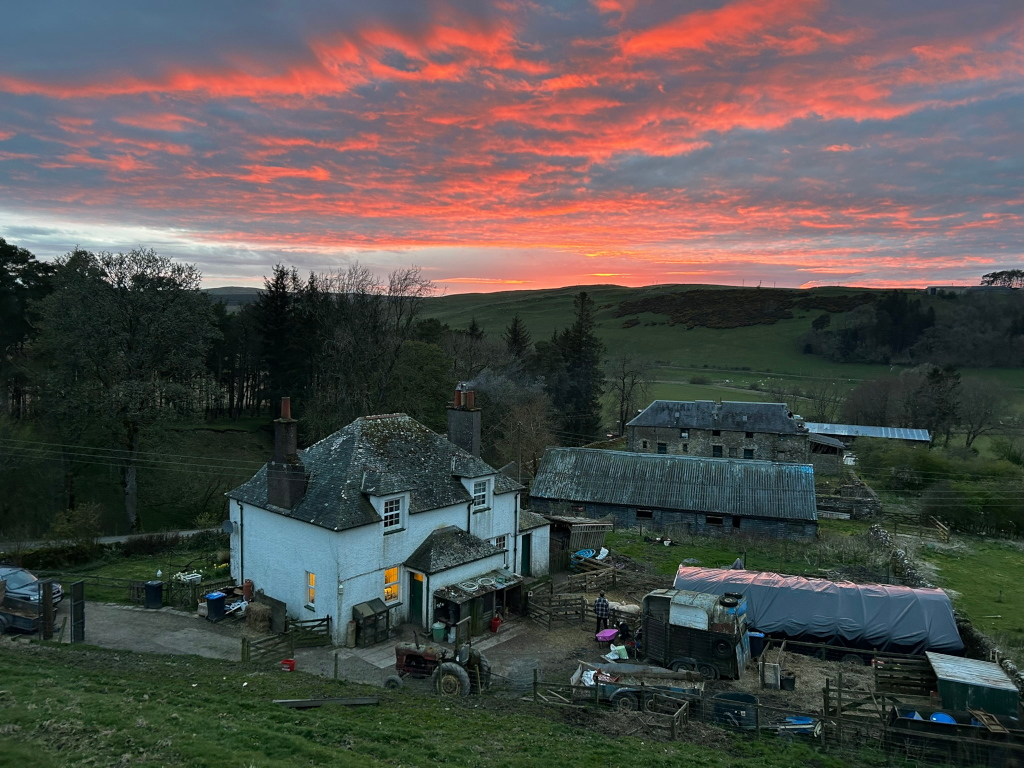 Rural scene at sunset. A white farmhouse, other farm buildings, and a covered trailer are in the foreground. Rolling green hills and a forest are visible in the background, all bathed in the warm, vibrant colours of a dramatic sunset sky. The overall impression is one of serene countryside tranquillity.
