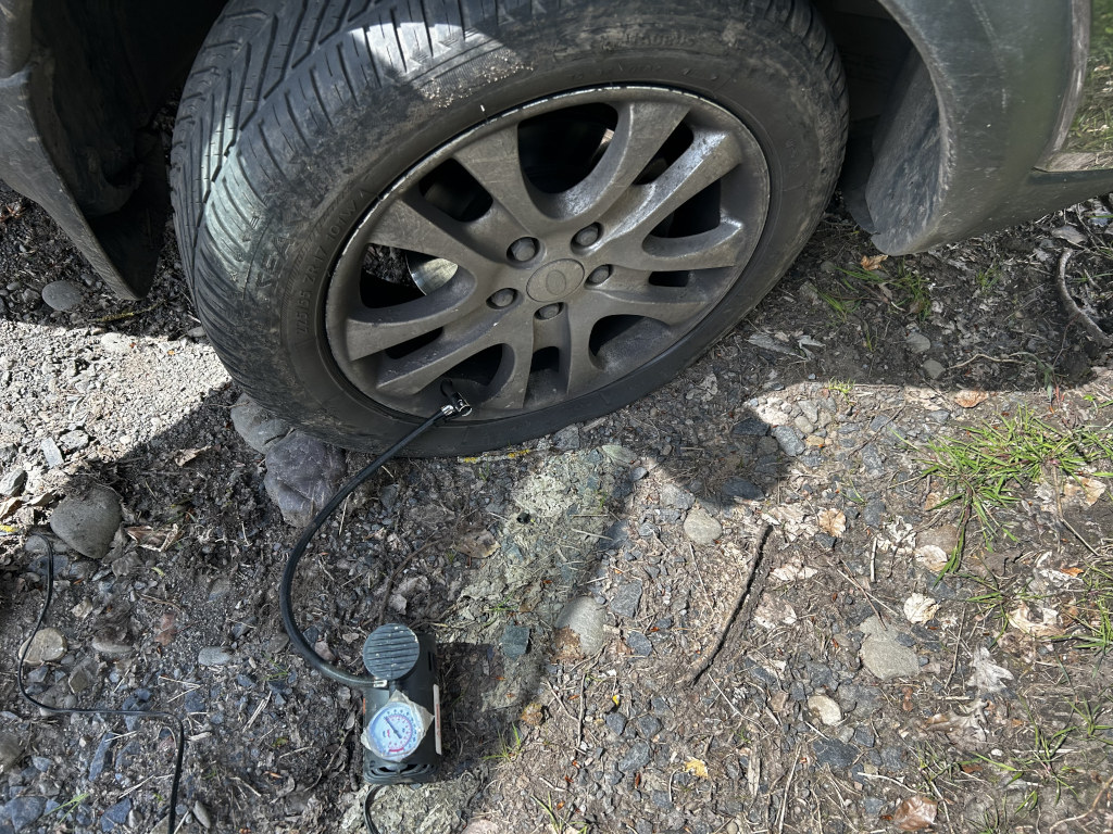 Car tire being inflated with a portable air compressor. The car is parked on a gravel surface. The focus is on the tire and the small, hand-held air pump connected to the tire valve.