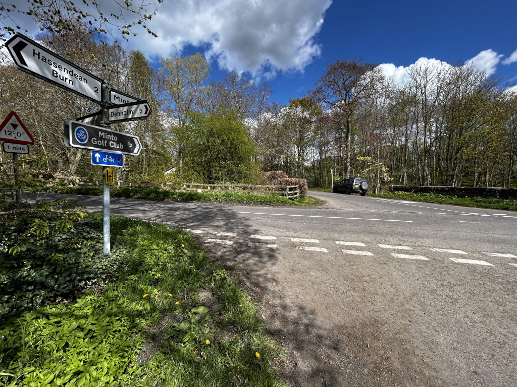 Rural road junction in Scotland on a sunny day. Several directional signs indicate the distances and directions to various locations, including Hassendean Burn, Minto, and Denholm. A dark-coloured SUV is parked near the junction, and the surrounding area is wooded with green undergrowth. The scene is peaceful and evokes a sense of quiet countryside.