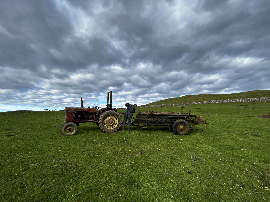 Farmer standing behind a vintage red tractor and trailer. The trailer is loaded with what appears to be hay or other farm produce. The scene is set in a rural landscape with a stone wall in the background, under a dramatic, cloudy sky. A sheep is visible in the distance. The overall impression is one of agricultural work in a remote and somewhat bleak but beautiful setting.