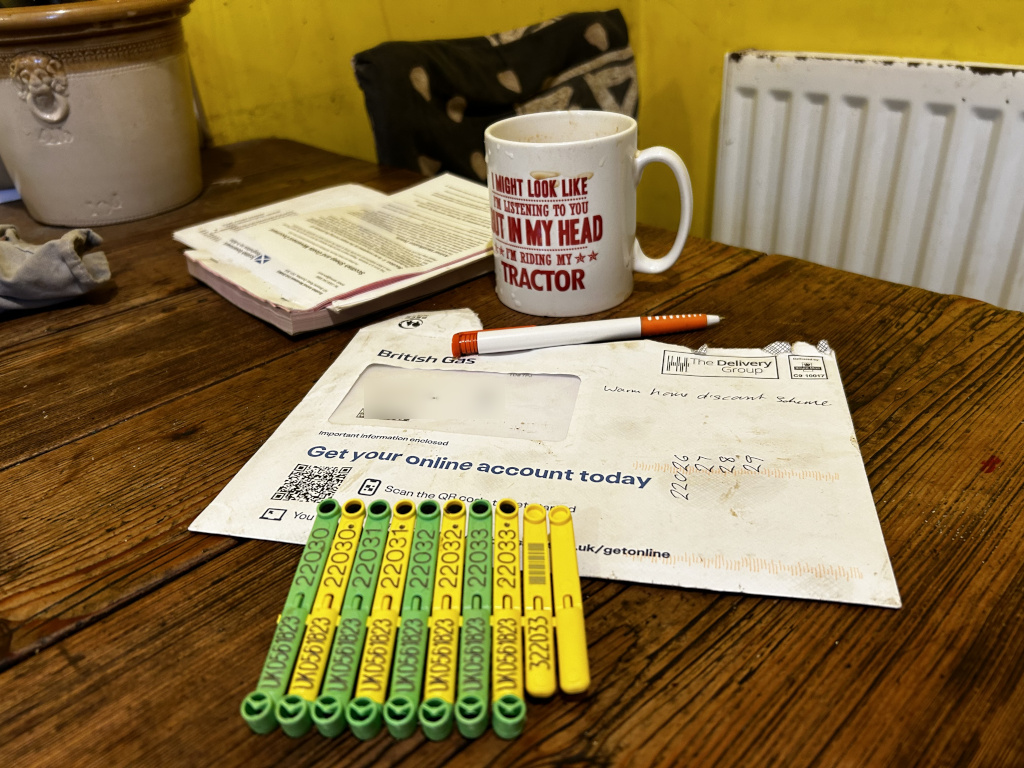 Wooden table with various items on it, seemingly in a domestic setting. The main focus is a British Gas envelope containing important information regarding setting up an online account. Several numbered gas meter tags are prominently displayed in the foreground. A mug with a tractor-related phrase implying a busy or distracted individual is present, along with some paperwork and a partially visible radiator. The overall impression is one of everyday life, possibly in a rural setting, interrupted by administrative tasks related to utility bills. The scene is casual and untidy, conveying a sense of normalcy and perhaps a brief pause in the day's activities.