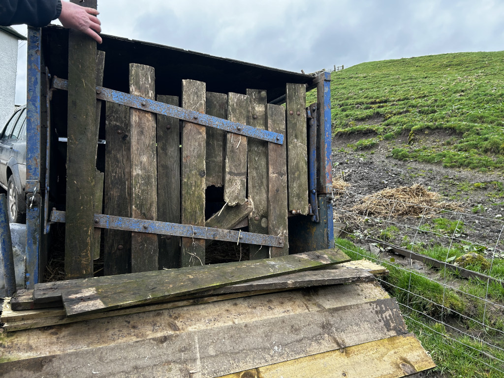 Dilapidated wooden structure, possibly a gate or part of a livestock trailer, with many of its wooden planks broken or missing. A person's hand is visible, appearing to be interacting with or inspecting the damaged structure. The setting is a rural landscape with a grassy hillside in the background.