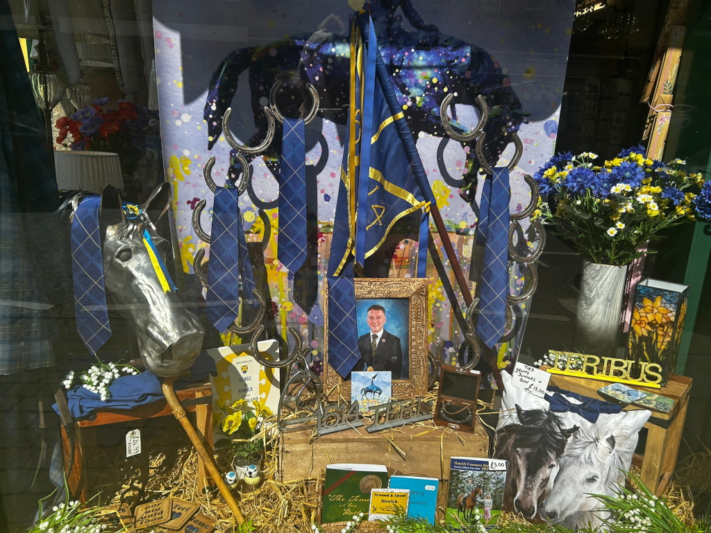 Window display, possibly for a shop selling equestrian-related items or gifts. The display features a metallic horse head, several blue ties hanging from a horseshoe rack, a framed photograph of a man in a suit, a blue and gold flag, flowers (including cornflowers), and various smaller items like books and coasters. The overall feel is one of tribute or commemoration, possibly related to a specific person or event. The colour scheme is predominantly blue and gold, with pops of yellow and white from the flowers. A pillow with a depiction of two horses is also visible.