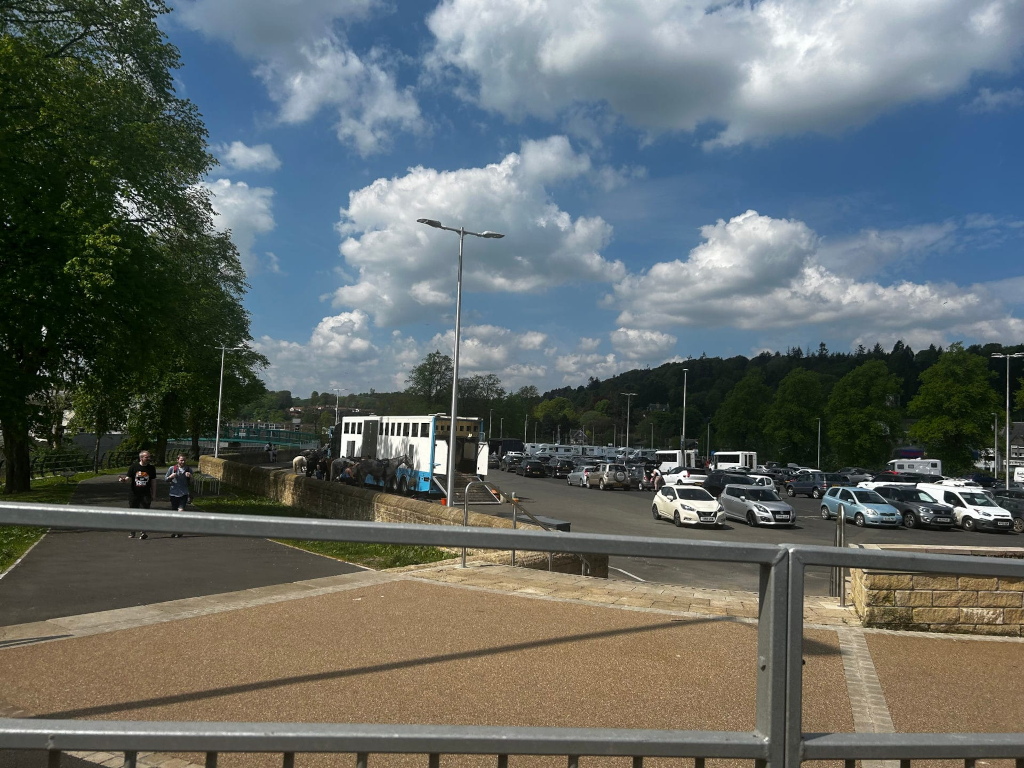 Sunny day with a partly cloudy sky. In the foreground is a paved area, possibly a park or walkway, with a metal railing.  In the background, a large number of cars are parked in a lot. Further back, a horse trailer is visible, suggesting perhaps a show or event. Two people are walking along a path in the lower left corner of the image. There's a lot of greenery, including trees and a hillside covered in vegetation, in the distance.