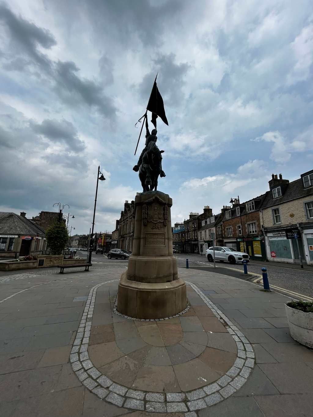 Statue of a person on horseback raising a flag, set atop a stone pedestal in a town square.  The statue appears to be a war memorial, with inscriptions visible on the pedestal. The background features a street with buildings characteristic of a small town or village. The sky is overcast.