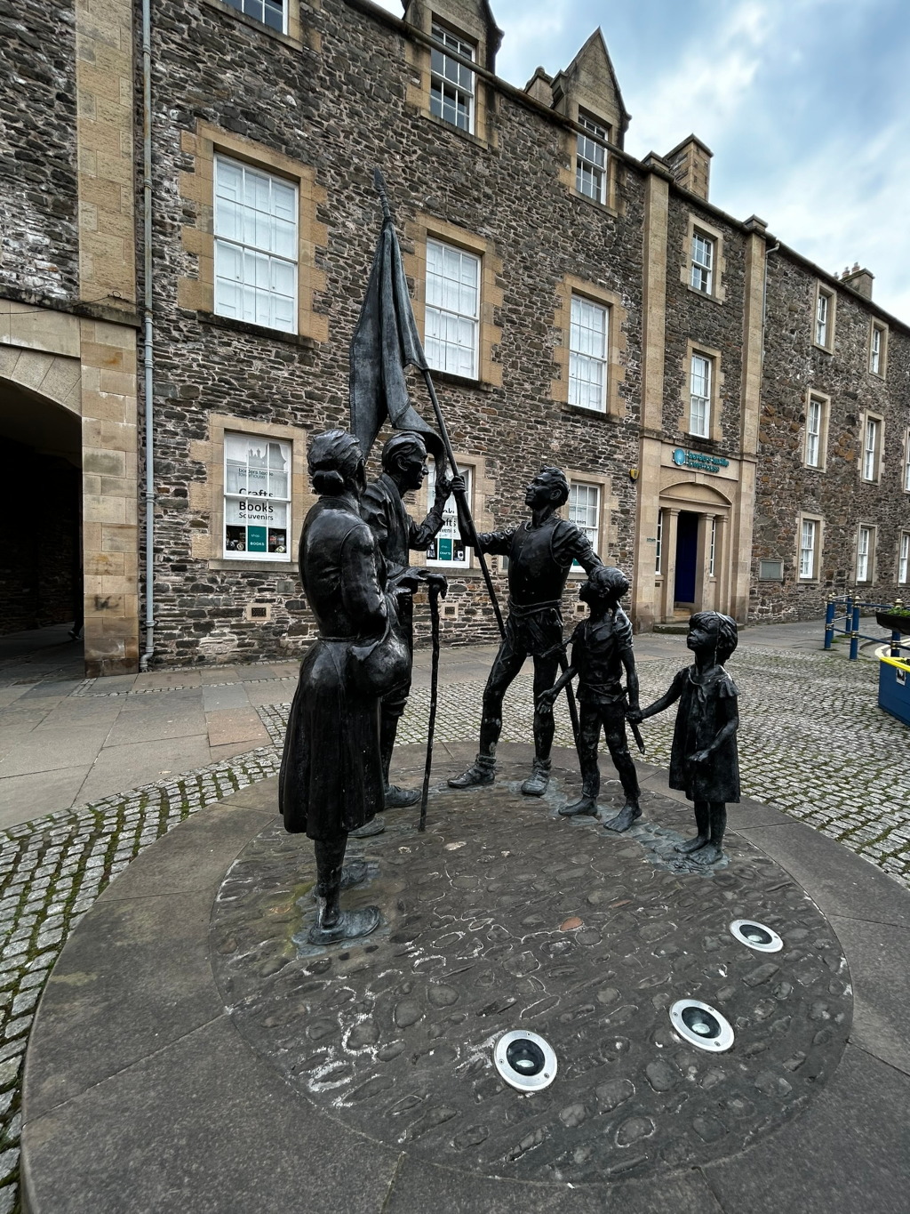 Bronze statue depicting a family of five—two adults, two children, and a baby—carrying a flag. The statue is located in a cobblestone plaza in front of a stone building. The scene suggests a journey or a pilgrimage. The somber tone of the sculpture, coupled with the overcast sky, evokes a sense of hardship and determination. The historical setting adds another layer of narrative, hinting at a moment of significant cultural or historical value.