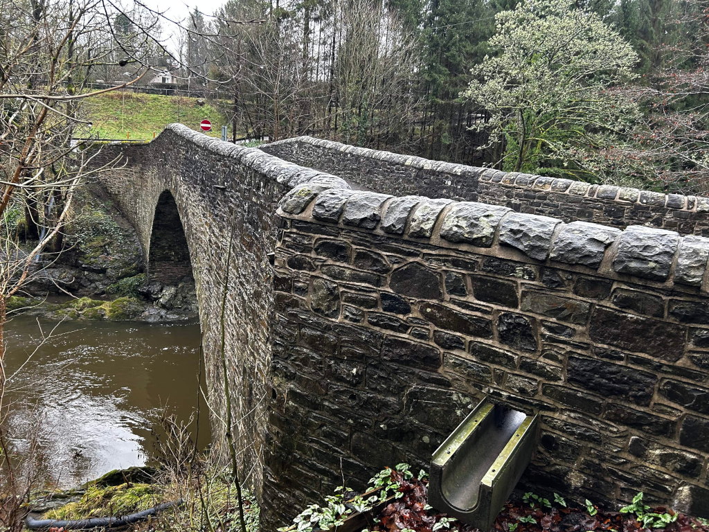 Stone arch bridge spanning a river. The bridge is made of dark grey stones, and the water below is brown. The surrounding area is wooded, with bare trees in the background suggesting it may be winter or early spring. A small, modern drainage pipe is visible at the base of the bridge's wall. A no entry sign can be seen in the distance on the bridge.