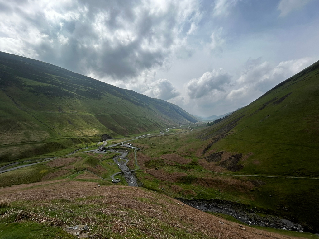 High-angle, long shot of a valley. Two steep, grassy hillsides converge in the middle distance, creating a V-shaped valley. A winding road and a small stream are visible at the bottom of the valley. The sky is mostly cloudy, with patches of blue showing through. The overall impression is one of wild, unspoiled natural beauty.