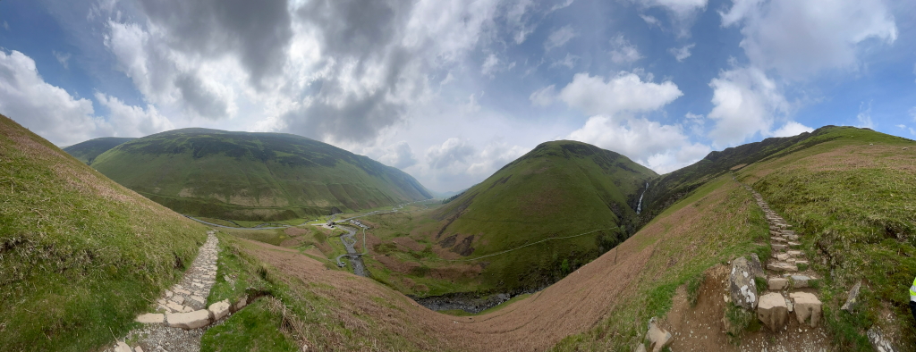 Panoramic view of a mountainous landscape. Rolling green hills, a valley with a small stream or river, and a stone pathway winding up one of the hillsides are prominent features. The sky is partly cloudy, with a mix of blue sky and fluffy white clouds. The overall impression is one of a serene, natural, and somewhat remote environment, possibly a hiking or walking trail in a rural area.