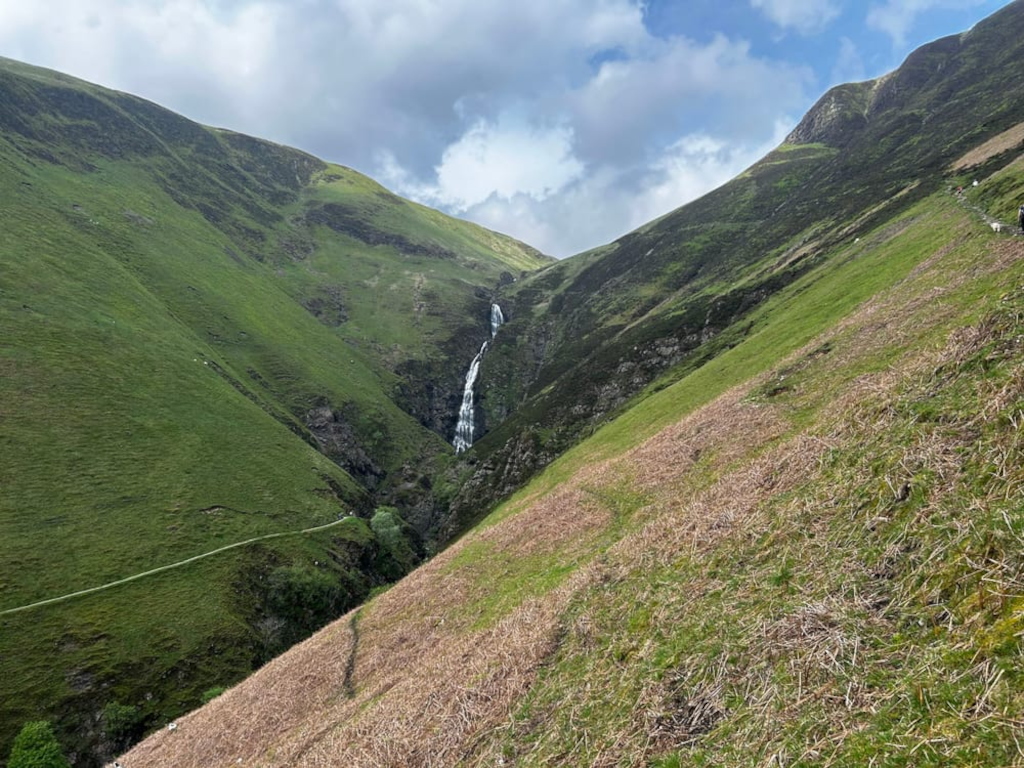 High-angle, long shot of a waterfall cascading down a steep, verdant valley nestled between two mountains. A faint trail is visible winding along one of the mountain slopes. The sky is partly cloudy, with patches of blue visible. The overall impression is one of wild, unspoiled natural beauty.