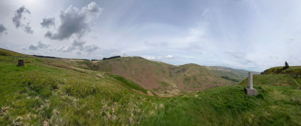 Panoramic view of a rolling green landscape, possibly in a mountainous region. The foreground is dominated by grassy hills, while the middle ground shows a valley or depression in the land, revealing a different coloured soil or vegetation. In the far distance, more hills and possibly mountains are visible under a partly cloudy sky. A stone marker or boundary post is noticeable in the lower right of the image, suggesting a point of interest or marking a boundary.