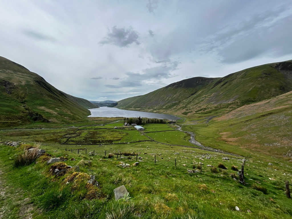 Tranquil valley scene, likely in a rural or mountainous region. A long, slender lake or reservoir stretches along the valley floor, nestled between steep, green hillsides. A small farm or homestead with a simple building is visible near the lake's edge, suggesting a secluded and peaceful life. Stone walls and fences indicate pastureland and possibly agricultural activity. The overall mood is serene and evokes a sense of isolation and the beauty of untouched nature. The sky is overcast, contributing to the calm, slightly moody atmosphere.