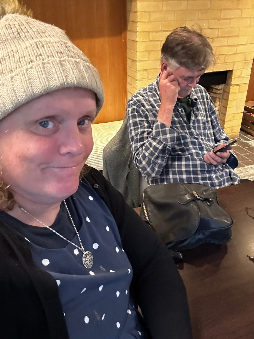 Leonie and Charlie seated at a dark brown table, possibly in a pub or restaurant setting. Leonie in the foreground is wearing a beige knit beanie, a navy blue polka-dot dress, and a black cardigan. She is wearing a silver necklace with a circular pendant and is looking directly at the camera with a slight smirk. Charie in the background is seated slightly behind and to the right, he's wearing a blue and white plaid shirt. Charlie is preoccupied, looking down at a mobile phone he's holding in his hands. He appears thoughtful or perhaps slightly distracted. A leather bag sits on the table between them. The background features a brick fireplace and wooden panelling, suggesting a rustic or traditional interior. The overall atmosphere is casual and somewhat intimate.