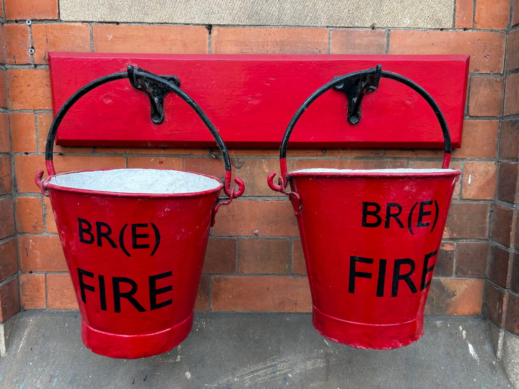 Two vintage red fire buckets hanging on a red wooden board affixed to a brick wall. The buckets are identical, marked with BR(E) FIRE in black lettering. They have black metal handles and appear to be relatively old, showing some wear and potentially some paint chipping. The brickwork suggests an older building, possibly a railway station or similar historical structure given the BR(E) marking, which likely stands for British Railways (Eastern Region). The overall impression is one of historical preservation.
