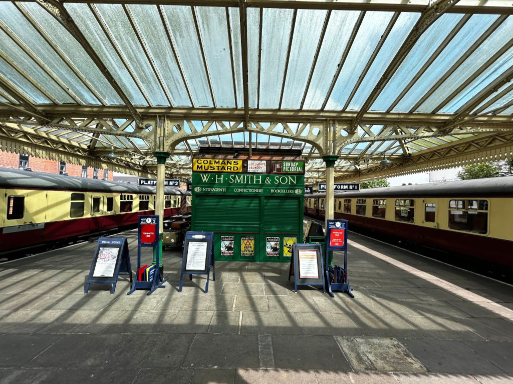 Vintage train station platform under a glass-roofed canopy. Two vintage railway carriages are parked on the platform. A small kiosk in the center displays advertisements for Colman's Mustard and W.H. Smith & Son, suggesting a period setting, possibly early to mid-20th century. Several A-frame advertising stands are also present. The scene is bathed in sunlight, casting shadows that accentuate the platform's architecture and the vintage aesthetic.