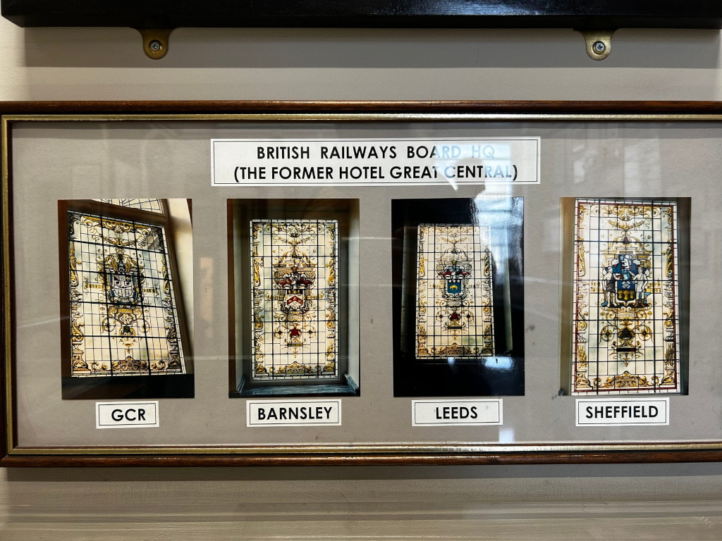Framed display showcasing four photographs of stained-glass windows. Each window features a coat of arms, and each photograph is labeled with a location: GCR, Barnsley, Leeds, and Sheffield. The display's title indicates that these windows were located in the British Railways Board Headquarters, formerly the Hotel Great Central. The image likely documents the historical significance of these windows and their connection to British railway history.