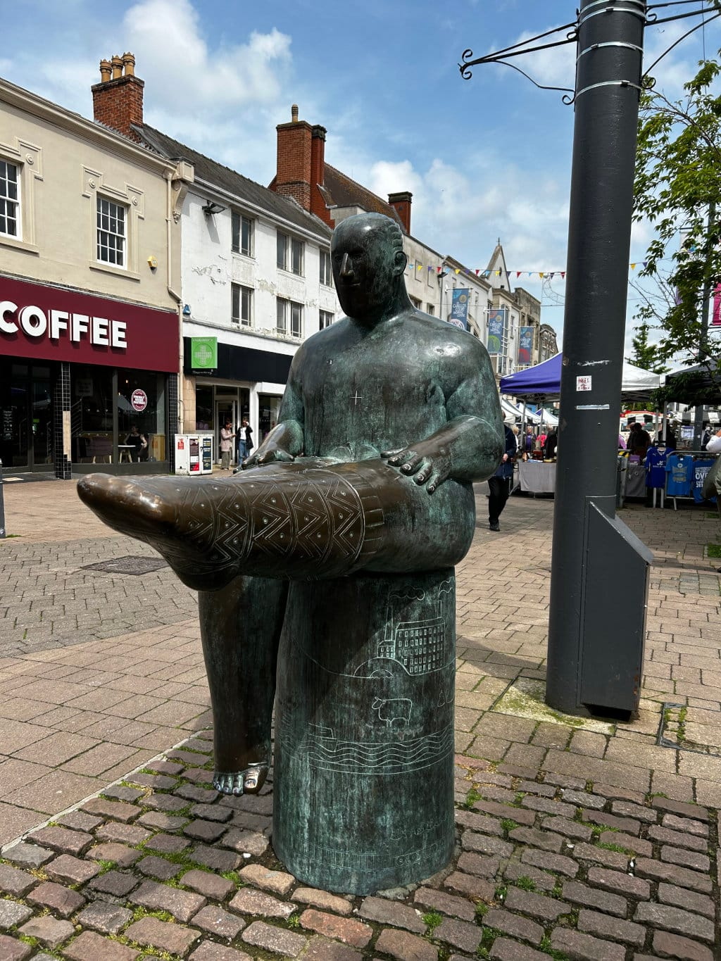 Bronze statue of a seated, robust figure holding a large, patterned stocking or sock. The statue is situated in a town square, with buildings and market stalls visible in the background. The base of the statue is decorated with etched designs depicting a landscape. The overall feel is one of public art in a bustling urban setting.