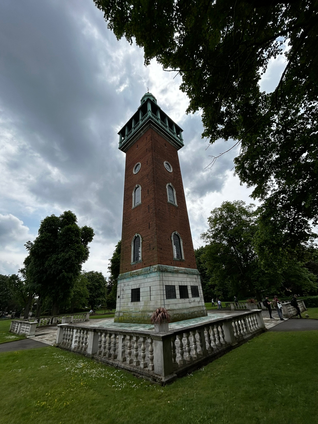 Tall, slender brick tower standing in a park. The tower is capped with a green-roofed cupola. It has several small windows and appears to be a clock tower or bandstand-type structure. The base of the tower is light-coloured stone, and a low stone balustrade surrounds the immediate area. Green trees and a cloudy sky form the backdrop. People are visible in the distance within the park. The overall impression is one of a somewhat aged, yet stately structure in a peaceful setting.
