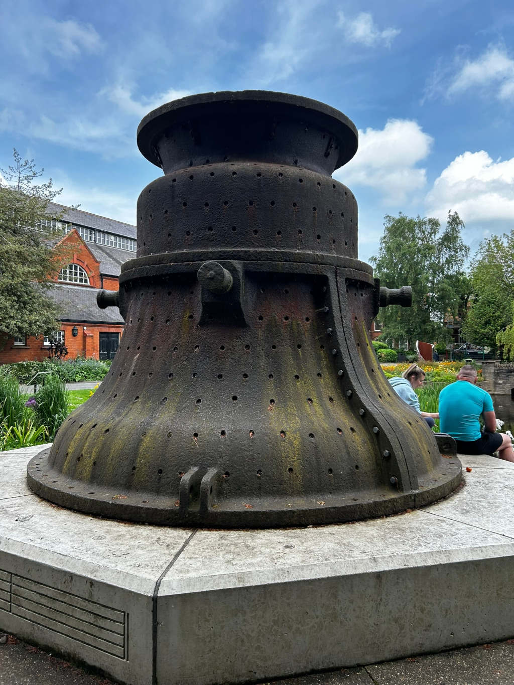 Large, dark-grey metal structure, possibly a piece of industrial machinery or a sculptural artwork, that resembles an oversized bell or a hopper. It's weathered and shows signs of age and rust, with numerous small holes throughout its surface. The structure sits on a light grey concrete base in a park-like setting, with a brick building and some greenery visible in the background. Two people are seated behind the structure, partially obscured from view. The overall impression is one of industrial history presented within a peaceful, modern environment.