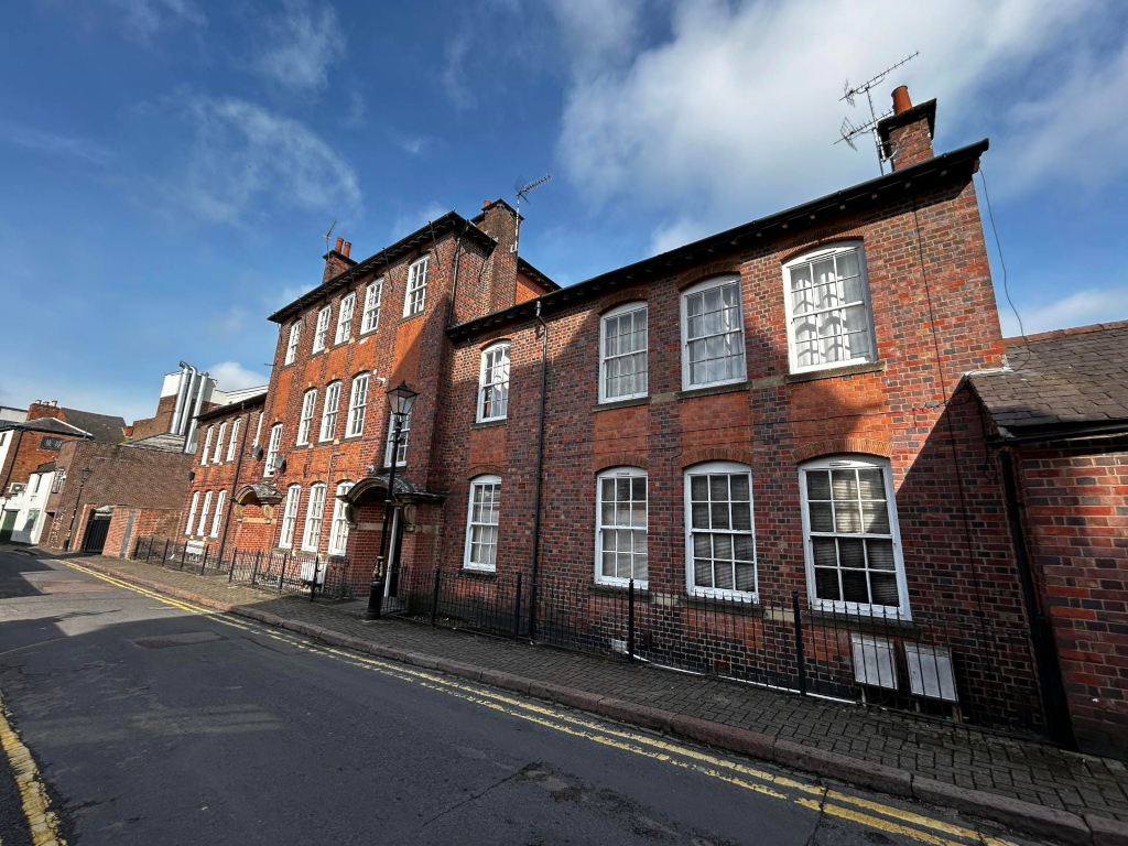Three-story brick building on a quiet street under a partly cloudy sky. The building is red brick with white-framed windows and appears to be residential, possibly converted from a larger structure. It has a slightly aged, historical look. The street is paved, and a small section of a neighboring building is visible. The overall scene is peaceful and somewhat quaint.