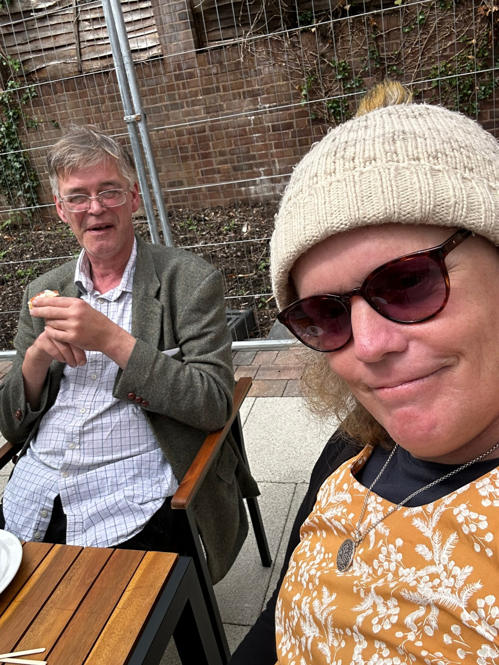 Charlie and Leonie sitting outdoors at a table. Charlie is wearing glasses and a jacket, and he's eating something. Leonie is wearing a knitted hat, sunglasses, and a mustard-coloured dress. They appear to be enjoying a casual meal together in a patio setting, partially enclosed by a metal fence. The background suggests a slightly urban or semi-rural outdoor space.