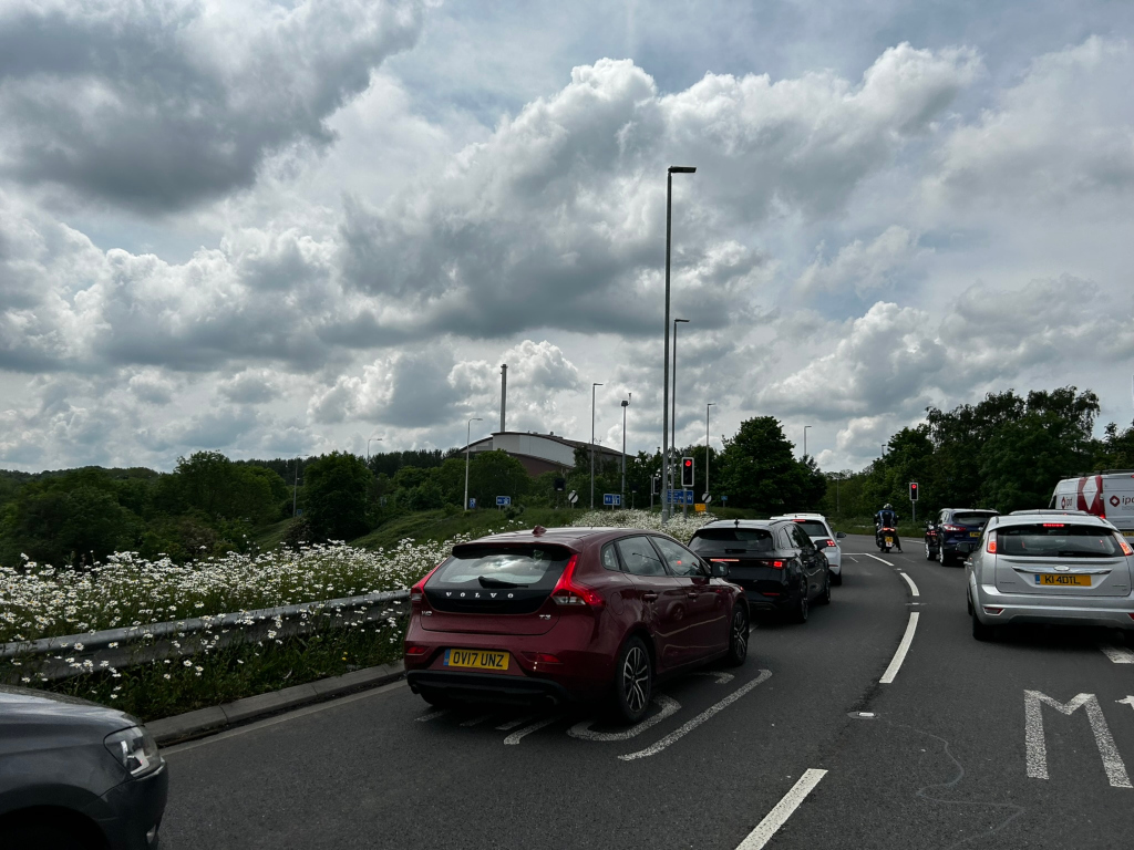 Traffic queue on a road, under a cloudy sky. In the foreground, there are several cars stopped or slowing down, with a burgundy Volvo in the centre. A field of white flowers runs along the side of the road. In the background, a large industrial or commercial building is visible beyond some trees. The overall scene suggests a moment captured during a typical day on a busy road, possibly near an industrial area.