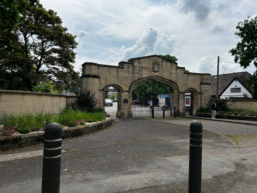 Stone archway, possibly the entrance to a park or institution. The archway features a coat of arms. Beyond the gates, there are buildings and a person walking. The overall style suggests a historical or traditional setting. Landscaping with flowers and plants is visible around the entrance. The sky is partly cloudy.