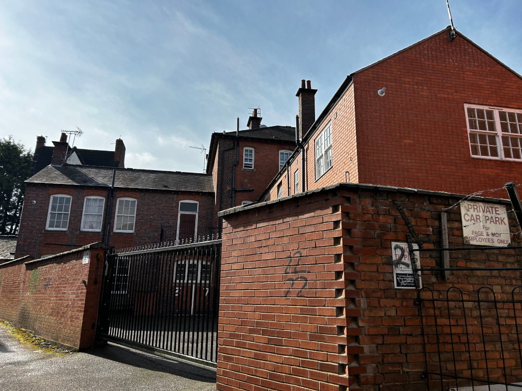 Back of a row of brick buildings. A black metal gate blocks access to a private car park, designated for employees only. The brickwork shows some age and wear, with graffiti visible on one wall. The overall impression is of a somewhat secluded, utilitarian space behind larger buildings. 