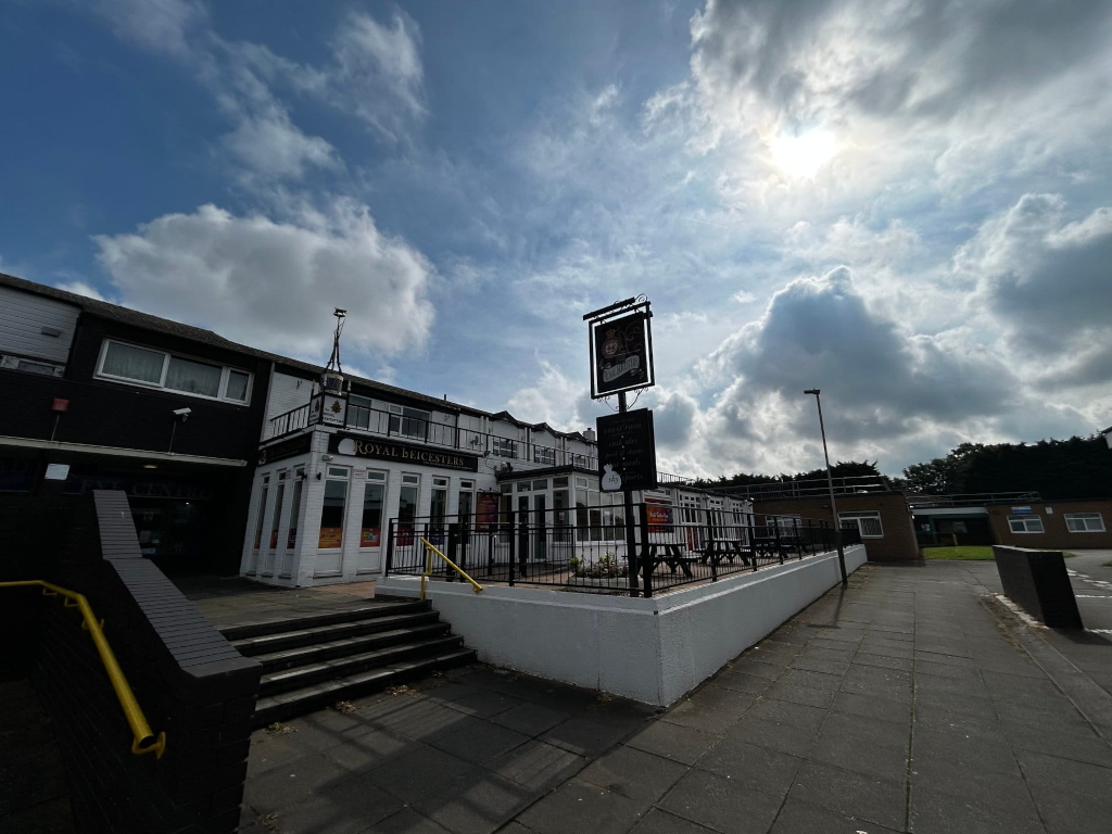 Exterior of a pub called Royal Beicasters. The building is predominantly white with black accents. There's a small patio area with tables and chairs enclosed by a low white wall and a black fence. The sky is partly sunny with large, puffy clouds. The overall feel is daytime, and the scene appears to be in a somewhat residential area.
