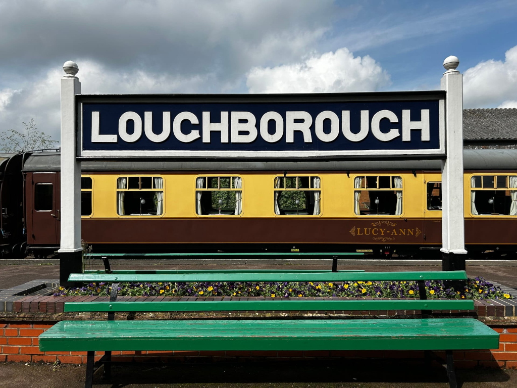 Railway station scene. A green park bench sits in the foreground, planted with colourful flowers. Behind it is a railway station sign proclaiming LOUGHBOROUGH. In the background, a portion of a vintage, yellow and brown passenger railway car, named LUCY-ANN, is visible. The overall setting suggests a charming and possibly historic railway station.