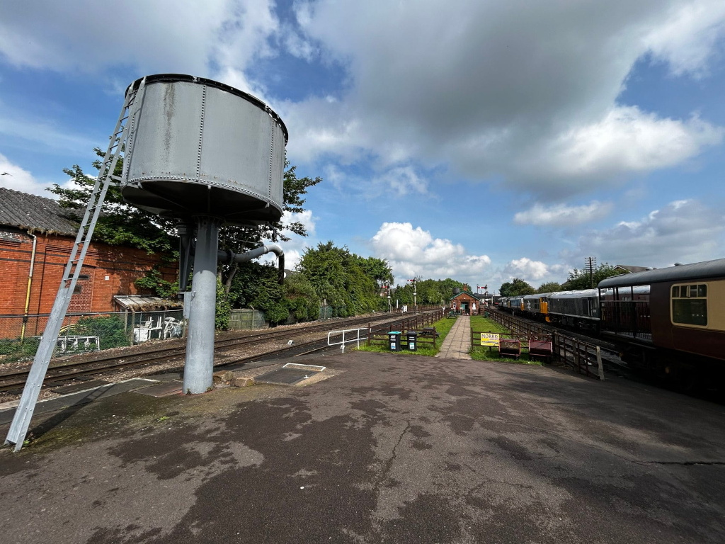 Railway scene on a partly cloudy day. In the foreground is a large, cylindrical water tower with a metal ladder attached, standing on a grey platform next to railway tracks. Beyond the water tower, a line of passenger carriages are visible, parked at a station with a small building and some greenery.