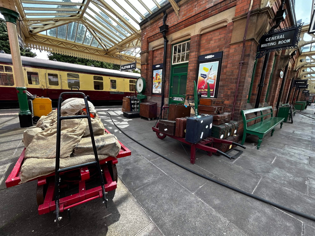 Vintage train station platform. A railway carriage is partially visible in the background. In the foreground, a red luggage trolley is laden with burlap sacks, and nearby, a collection of vintage suitcases sits on a smaller, similar trolley. A green bench is visible along the brick station building, which features vintage travel posters. The overall scene evokes a nostalgic atmosphere, possibly suggesting a film set or historical reenactment.