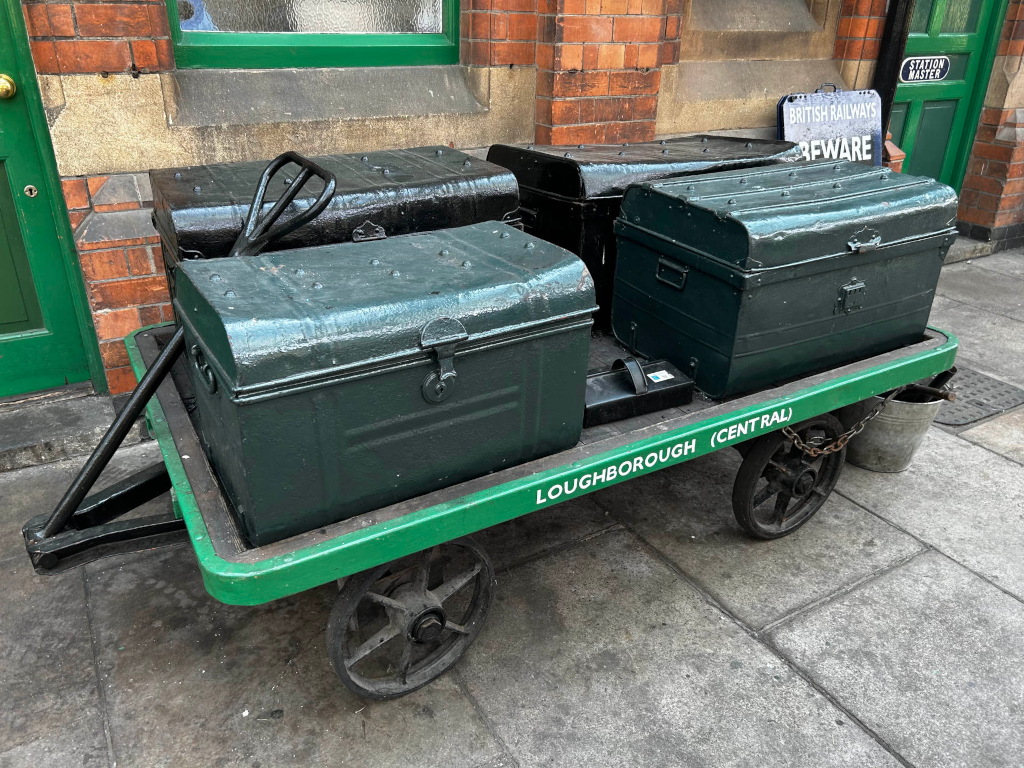 Green luggage trolley, labelled Loughborough (Central), carrying several dark green metal trunks. The trolley is parked outside a brick building with a green door and window. A small metal bucket sits next to the trolley. A Beware sign is visible on the building.