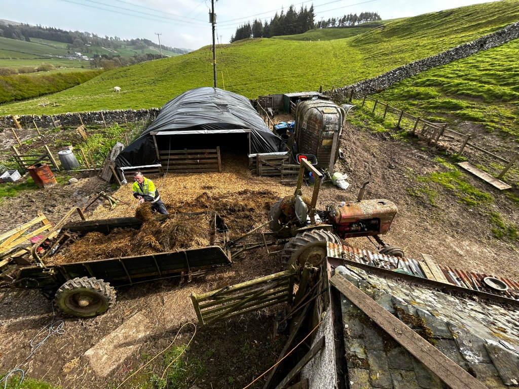 High-angle view of a rural farm scene. Charlie is seen in the process of loading hay or straw into a trailer using a small tractor. The setting includes a tarp-covered structure possibly for animal shelter, another older trailer-like structure, and a stone wall. The background shows rolling hills typical of a rural landscape. The overall impression is one of farm work and a simple, rustic setting.