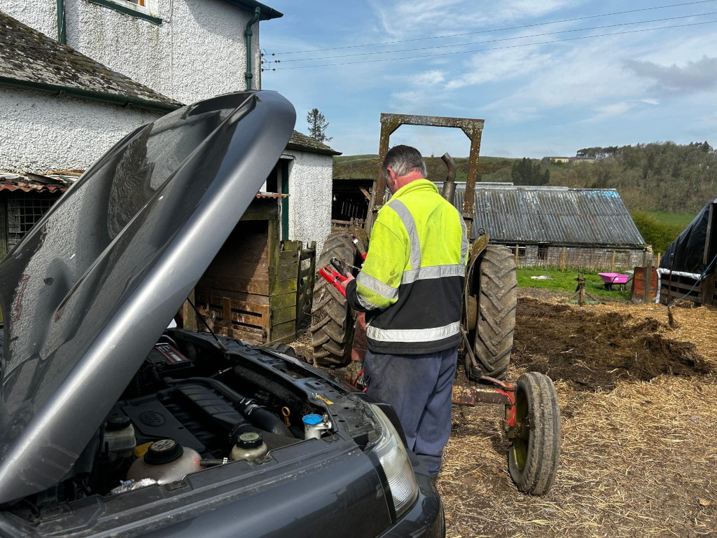 Charlie in a yellow high-visibility jacket standing behind a vintage tractor. He appears to be working on a dark grey vehicle with the hood open, possibly jump-starting it using jumper cables (partially visible). The setting is a farm, with bales of hay, a barn and other farm buildings in the background. The overall impression suggests a rural scene of everyday farm maintenance or repair.